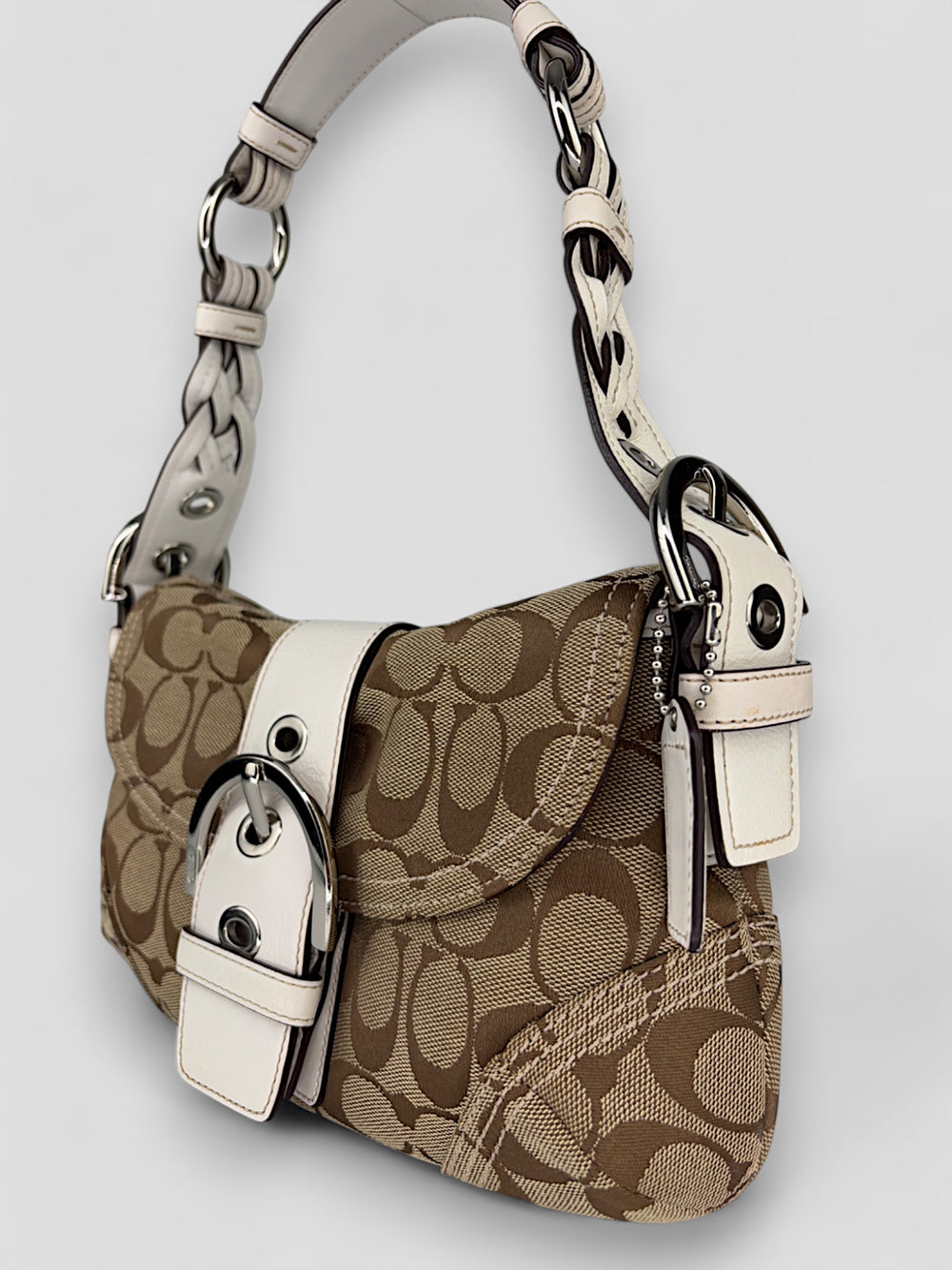 Coach Signature Strapbag mit beigem und braunem Logo-Muster, cremeweißen Lederdetails, silberfarbener Schnalle und geflochtenem Schulterriemen. Kompakt und stilvoll