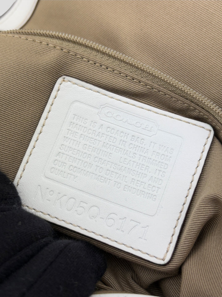 Coach Signature Strapbag mit beigem und braunem Logo-Muster, cremeweißen Lederdetails, silberfarbener Schnalle und geflochtenem Schulterriemen. Kompakt und stilvoll